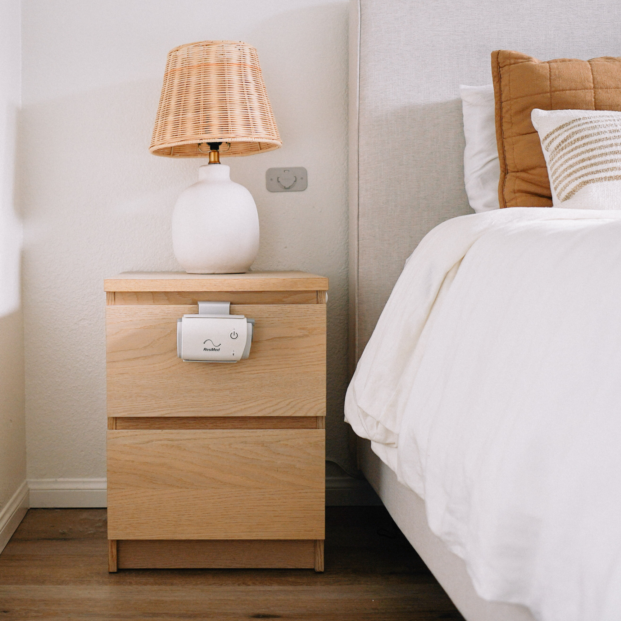 A bedside table in view with a bedlamp on top of it and the ResMed AirMini Travel CPAP Machine hooked onto a drawer